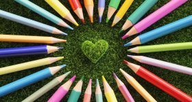 Colorful colored pencils radiate around a heart shape on green grass, symbolizing creativity and unity.